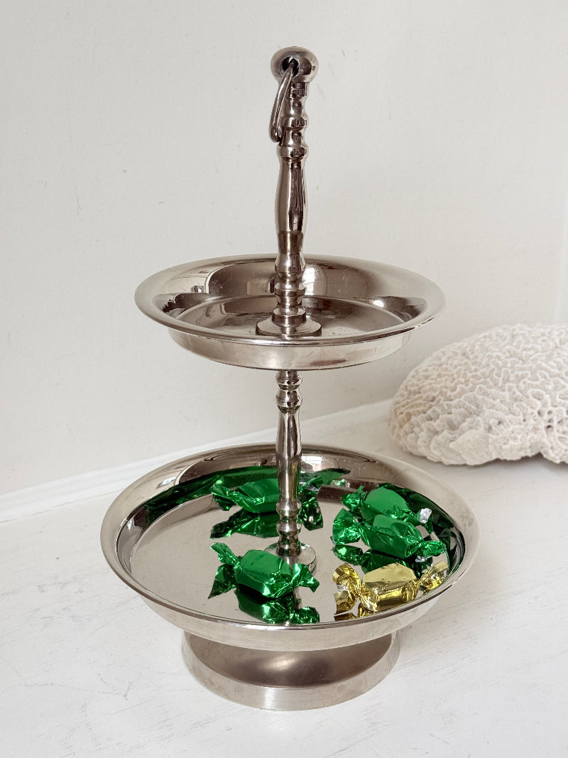 Two tier silver tone cake stand with polished reflective finish styled with green wrapped sweets on a white surface