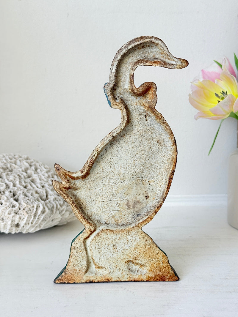 A rustic, vintage-style metal duck-shaped object with a distressed, weathered finish in beige, brown, and muted blue-green tones, displayed on a white surface with a white wall background and decorative props.