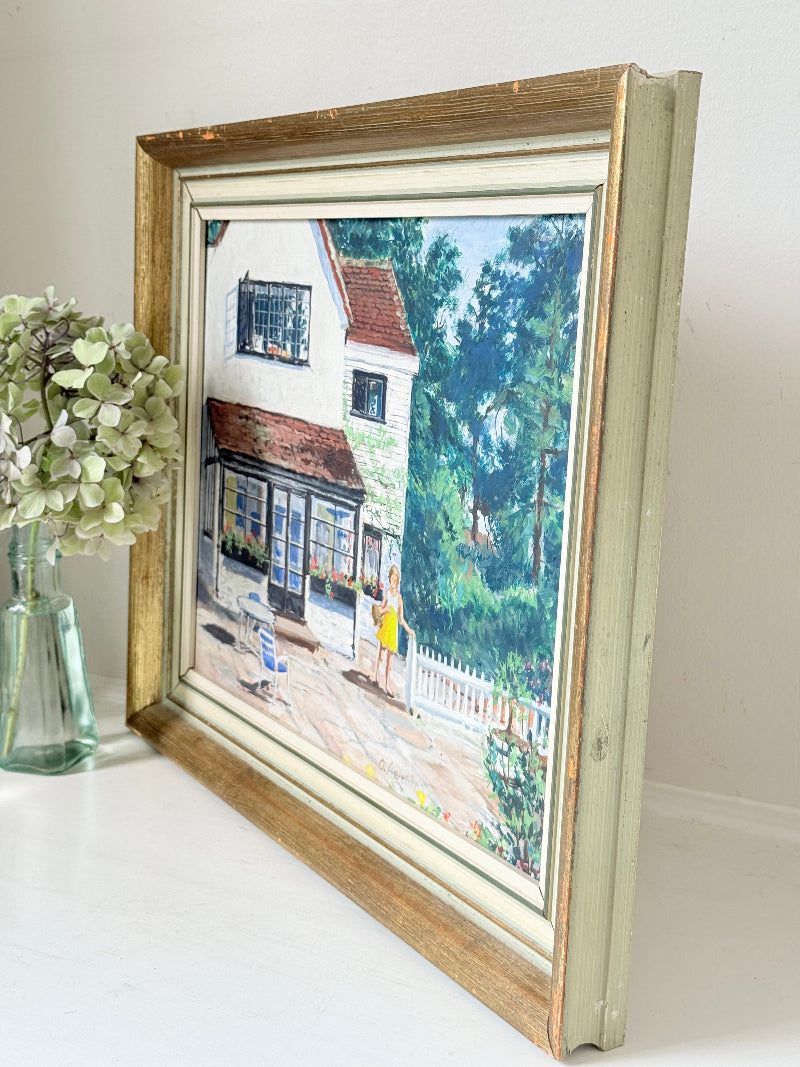 Oil on board painting of a girl in a yellow dress outside a cottage with picket fence, framed, photographed on a white surface