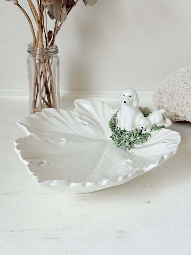 Italian ceramic dish featuring two white dogs on a sculpted leaf base, photographed on a white surface.