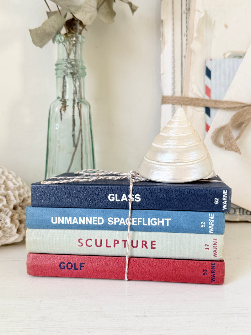 A stack of five hardcover books tied with twine, placed on a white surface with decorative props including a glass bottle, dried leaves, and a white cone-shaped object. The book spines read GLASS, UNMANNED SPACEFLIGHT, SCULPTURE, GOLF, and WARINE.