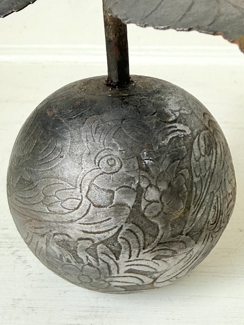 A close-up of a round, dark gray metal ornament or decorative piece with engraved floral and bird-like patterns, mounted on a vertical stem against a light off-white background.