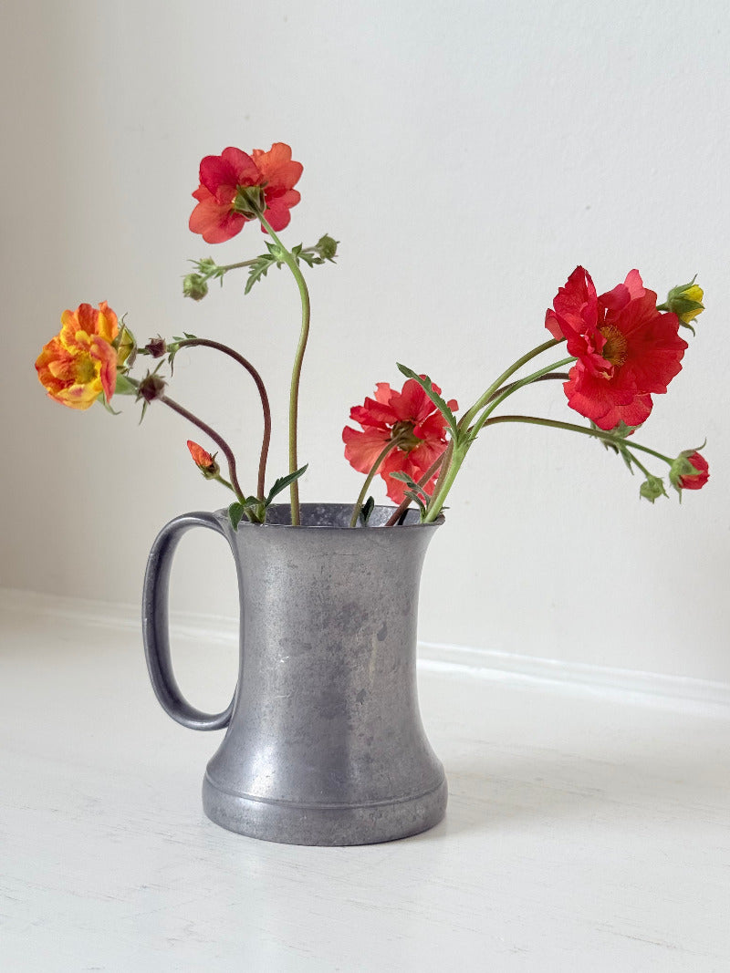 vintage pewter tankard with crown and rose mark and handle, styled with red and orange flowers on white surface