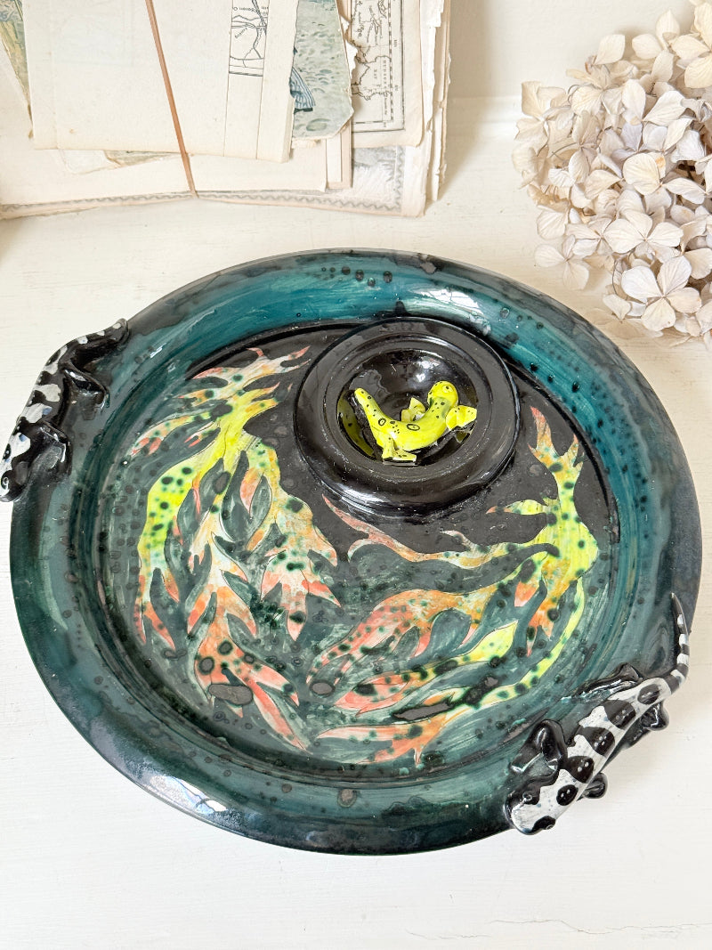 A handmade ceramic lidded pot with a matching saucer, decorated with a glossy teal glaze, hand-painted koi fish in yellow, orange, and green, and black-and-white dragon handles on the sides.