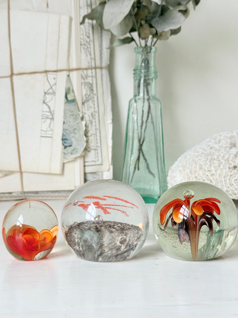 Trio of orange flower glass paperweights with green stems and controlled bubbles, photographed on a white surface