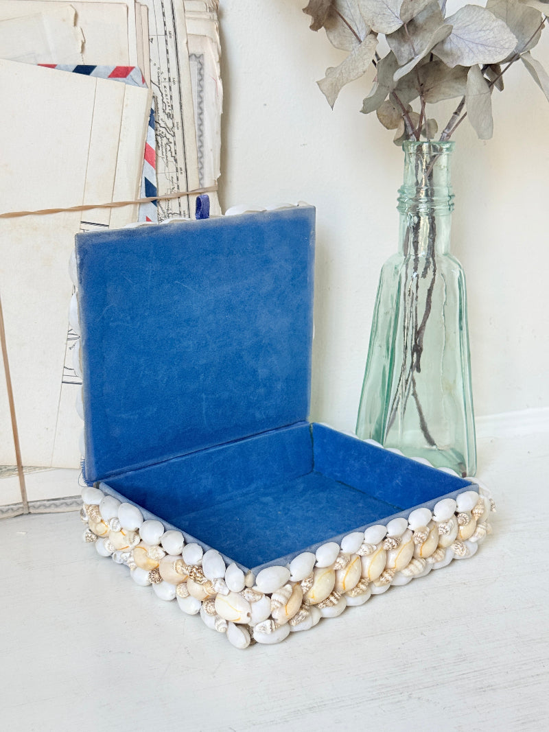 A square velvet box in a rich royal blue color, featuring a decorative border of white and gold seashells around the base. The box is shown open, styled on a white surface next to a vintage glass bottle vase with dried leaves.