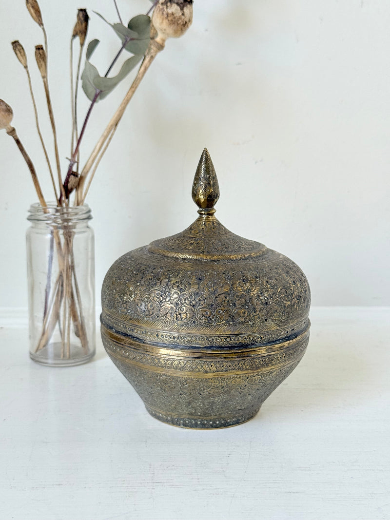 Indian Benares brass lidded jar with pointed finial and darkened interior, photographed on a white surface