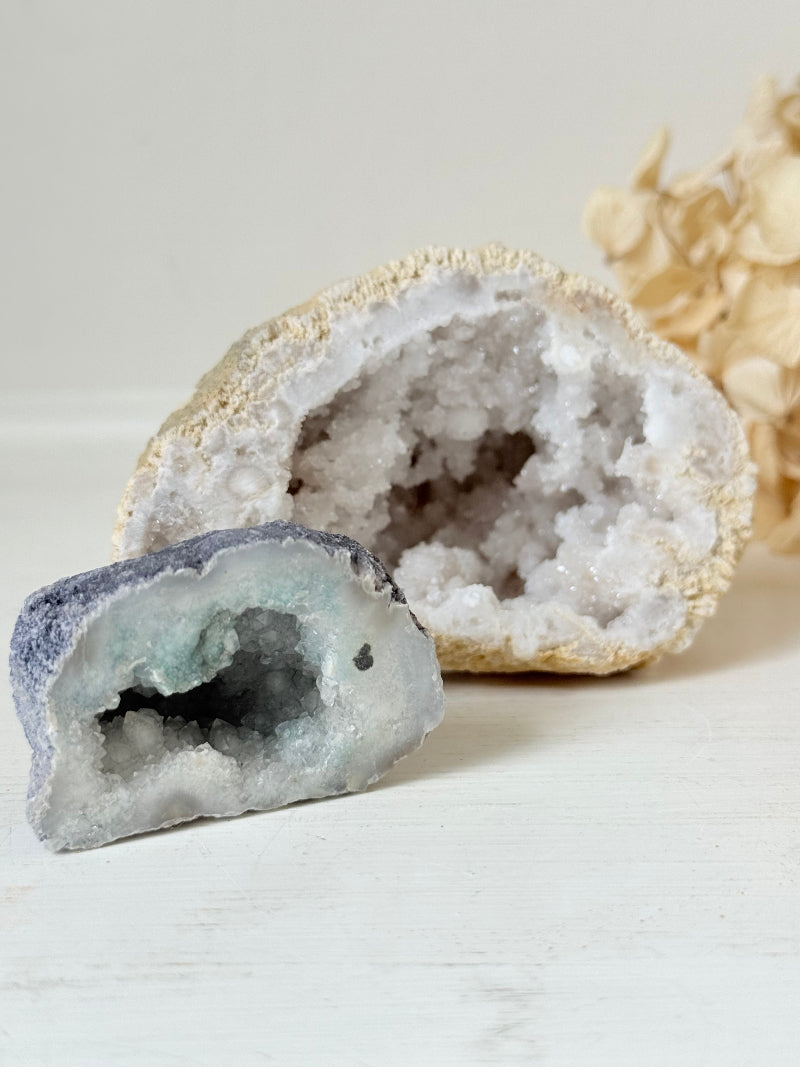 Pair of natural quartz geodes, one white and one lilac-toned, styled on a white surface with dried hydrangea