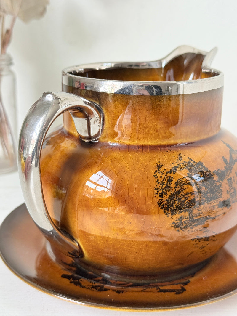 Antique Ridgways stoneware creamer jug and saucer photographed on a white surface in amber treacle glaze with black coaching scenes and silver lustre rim