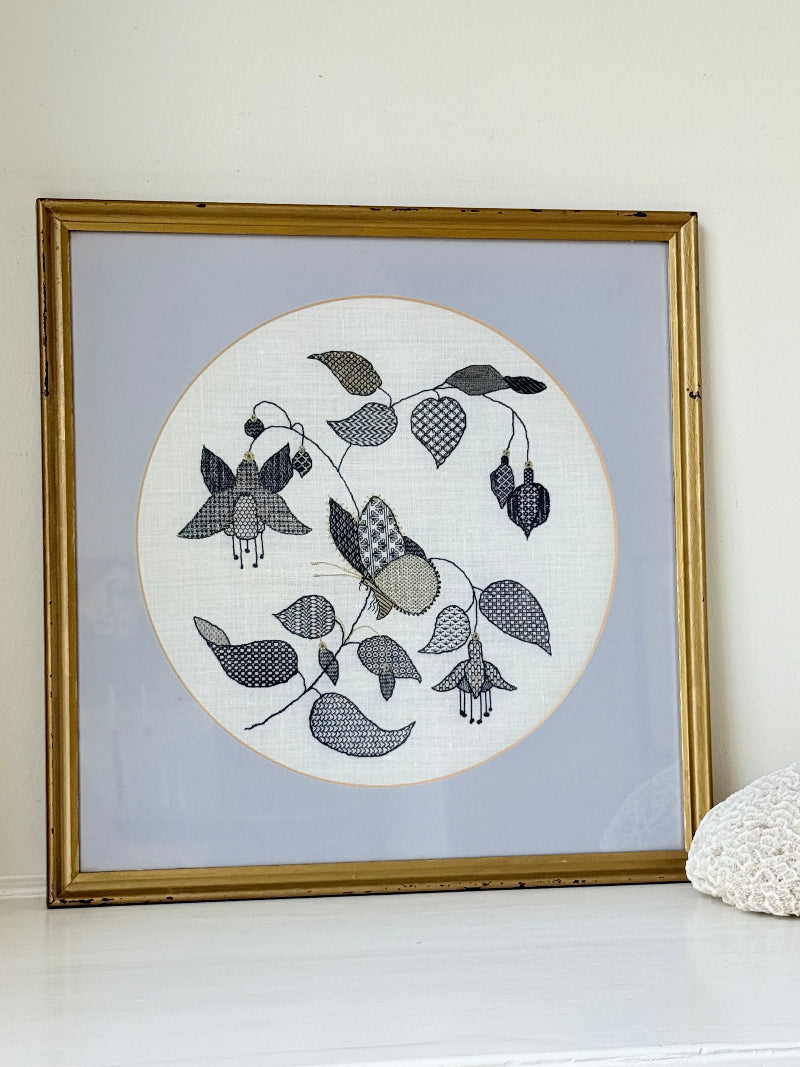 Framed circular hand embroidery of birds and fuchsia flowers in black and grey on white linen, set in gold frame, white surface background.
