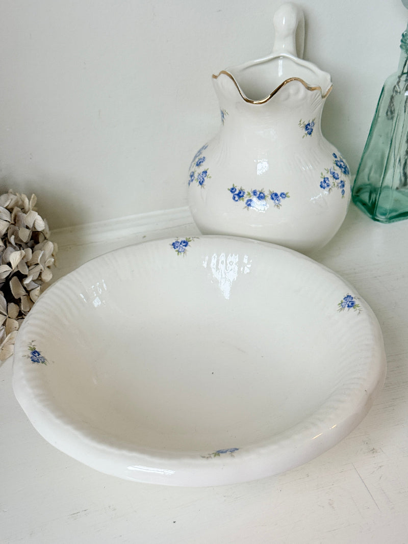 A white ceramic bowl with a fluted rim in the foreground and a matching white pitcher with blue floral patterns and gold trim in the background, set against a white wall.