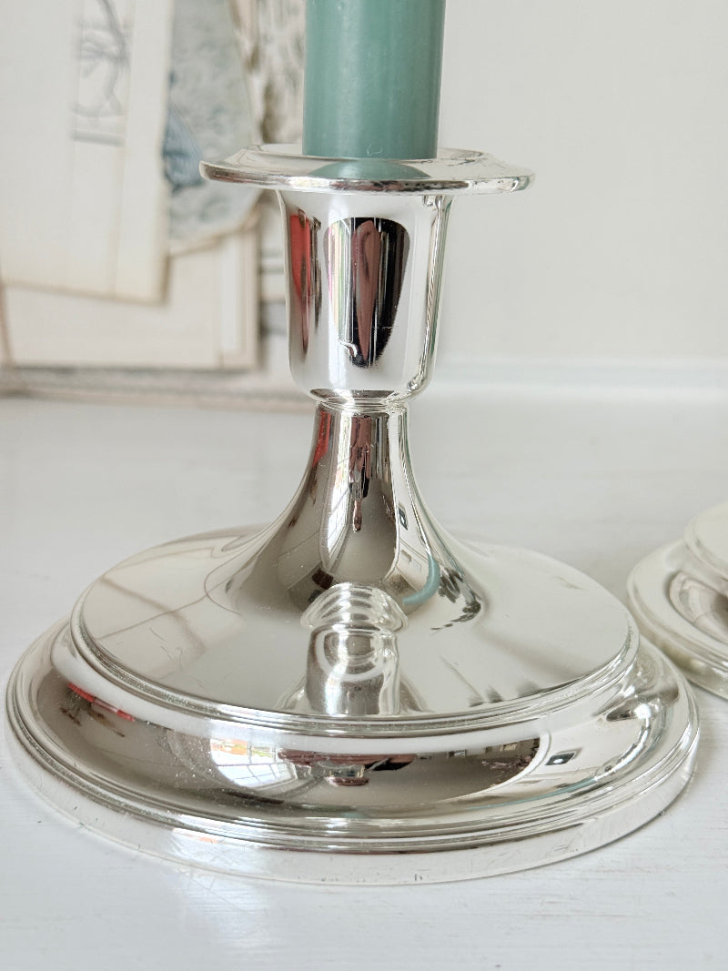 Pair of silver tone candlesticks with wide round stepped bases holding green candles, photographed on a white surface