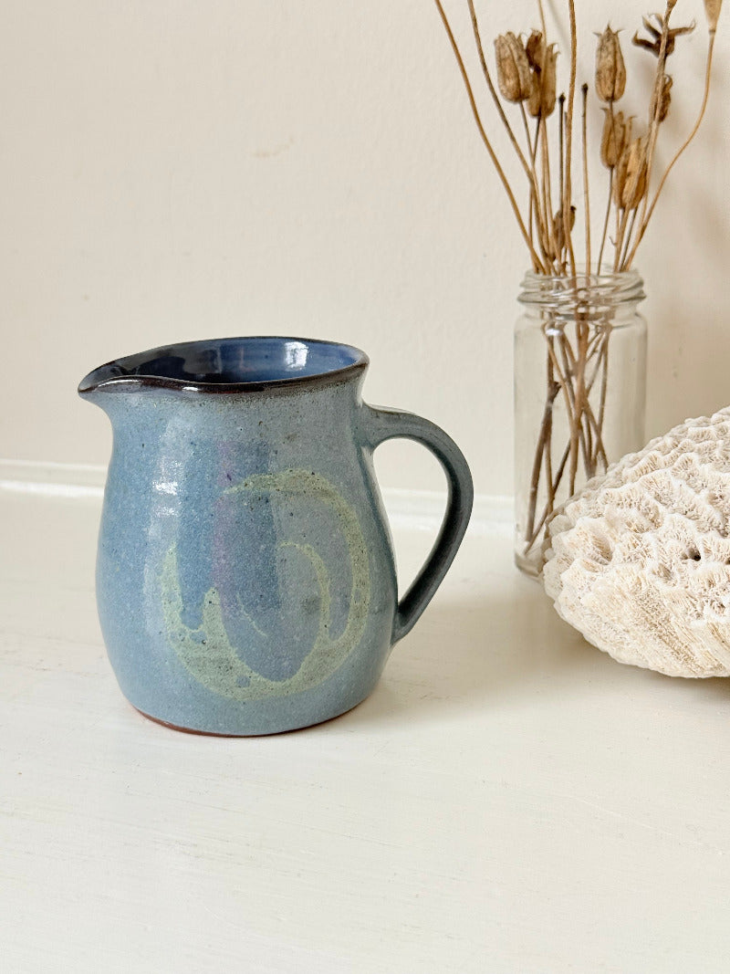 Vintage studio pottery milk jug with blue and green glaze, handmade with a rounded form and sturdy handle, styled on a white surface.