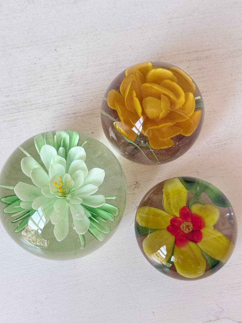 Trio of mixed flower glass paperweights in green, yellow and red, graduated in size, photographed on a white surface