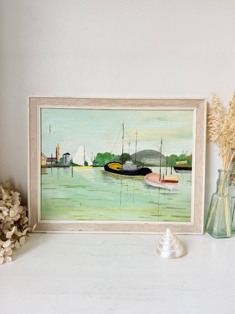 A framed watercolor painting of a harbor scene with boats and buildings, displayed on a white surface against a white wall with decorative dried flowers and a glass vase.