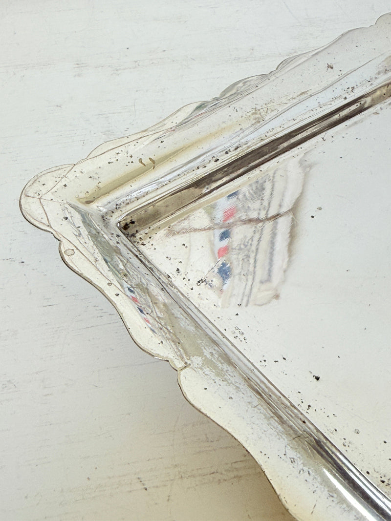 A vintage silver-toned metal tray with a scalloped edge, showing signs of age, wear, and rust, resting on a distressed white painted wooden surface.