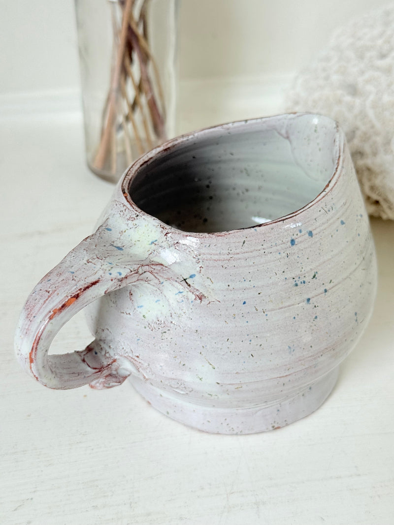 Studio pottery milk jug with soft pink and grey speckled glaze, photographed on a white surface