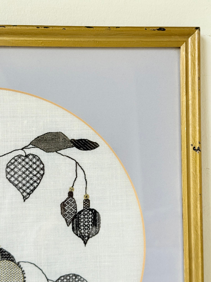 Framed circular hand embroidery of birds and fuchsia flowers in black and grey on white linen, set in gold frame, white surface background.