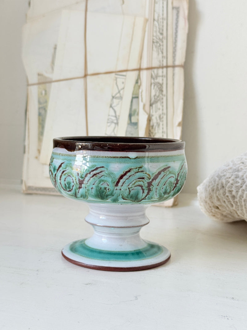 Hand thrown studio pottery footed bowl with green and brown swirling decoration photographed on a white surface