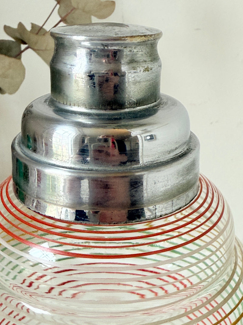 Vintage glass cocktail shaker with multicoloured stripes and silver lid photographed on a white surface.