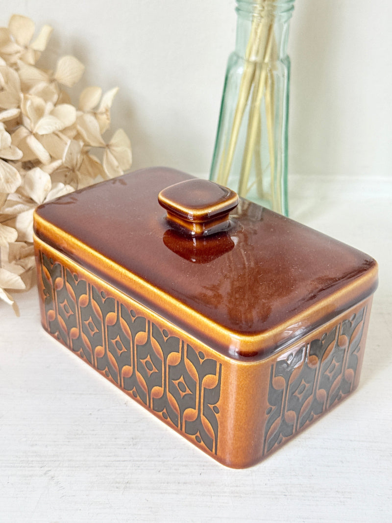 A rectangular ceramic butter dish with a matching lid, featuring a glossy brown and amber-brown patterned glaze, styled on a white tabletop with dried flowers and a glass reed diffuser in the background.