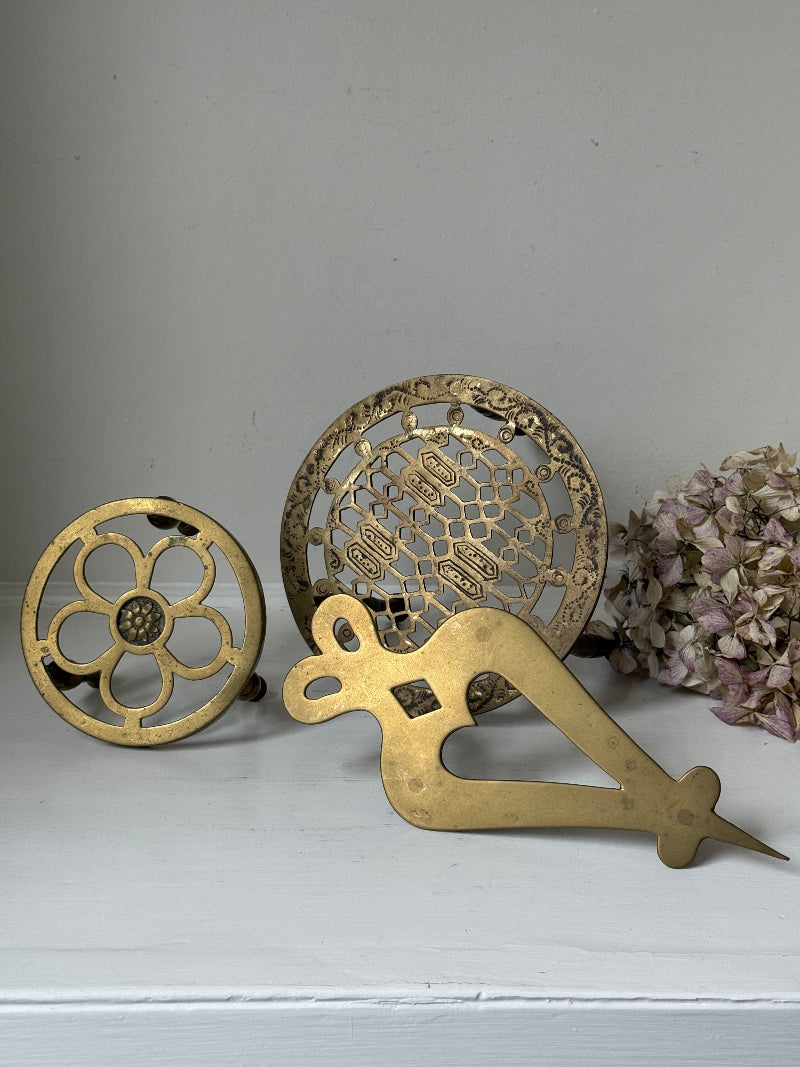Vintage brass trivet trio with ornate cutwork designs and aged patina, photographed on a white surface