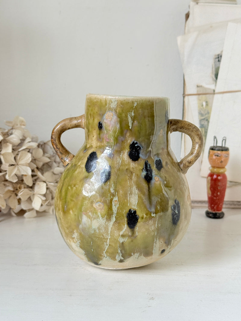 A handmade ceramic vase with a rounded body, two side handles, and a textured glaze in olive green, beige, and black speckles, displayed on a white surface with dried flowers and a small wooden doll in the background.
