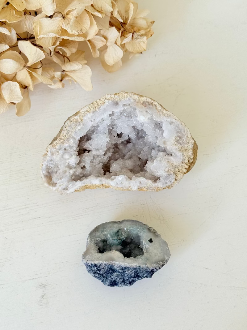 Pair of natural quartz geodes, one white and one lilac-toned, styled on a white surface with dried hydrangea