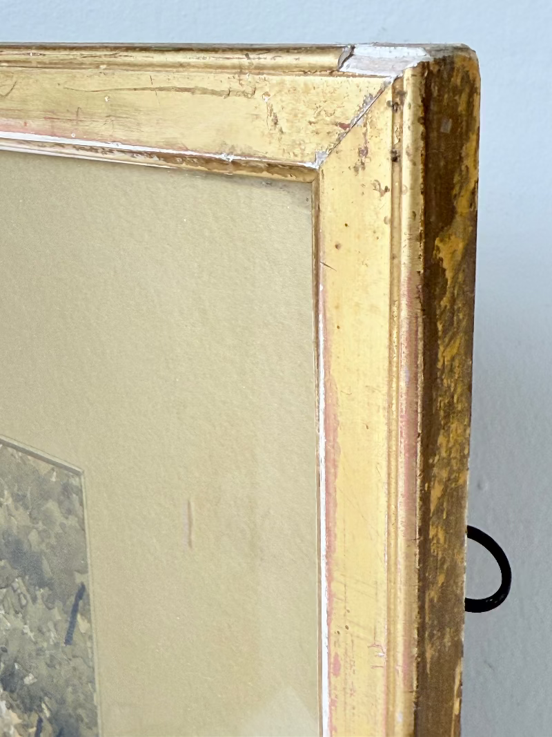 Antique framed watercolour of English village street with thatched cottages and figures, in gilt frame with wide mount, on white surface.