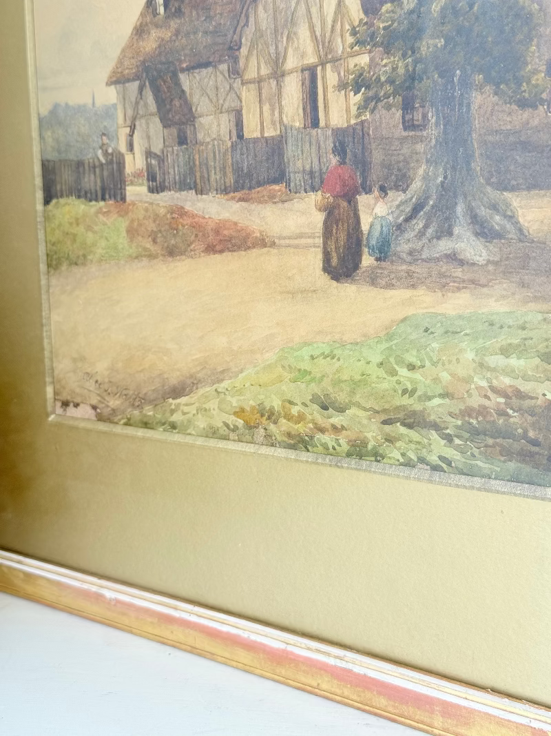 Antique framed watercolour of English village street with thatched cottages and figures, in gilt frame with wide mount, on white surface.