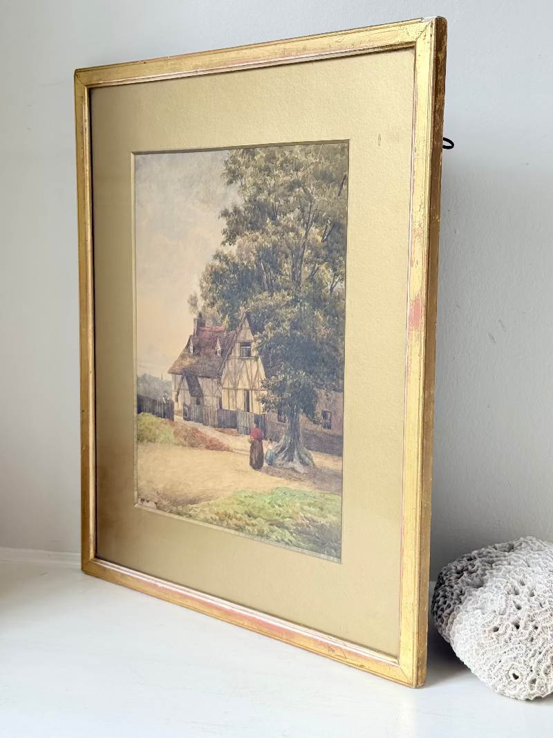Antique framed watercolour of English village street with thatched cottages and figures, in gilt frame with wide mount, on white surface.