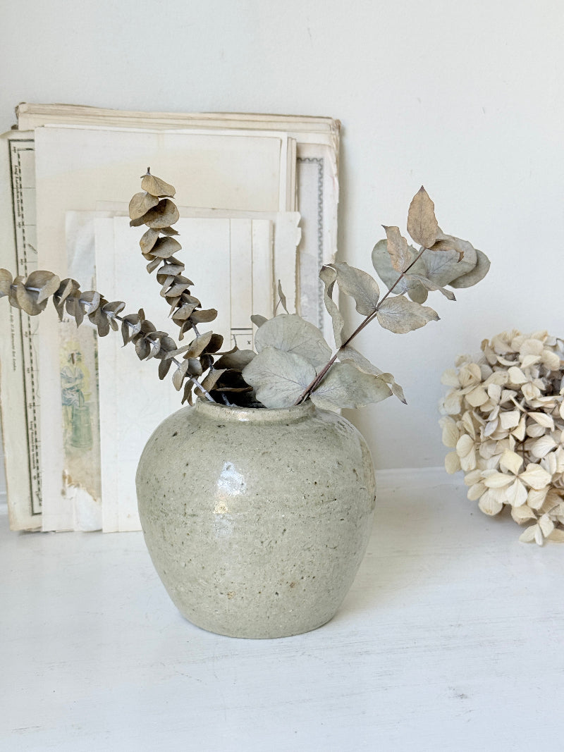 A speckled ceramic vase in a light gray-green glaze, styled with dried eucalyptus branches and a dried hydrangea on a white surface against a white wall.