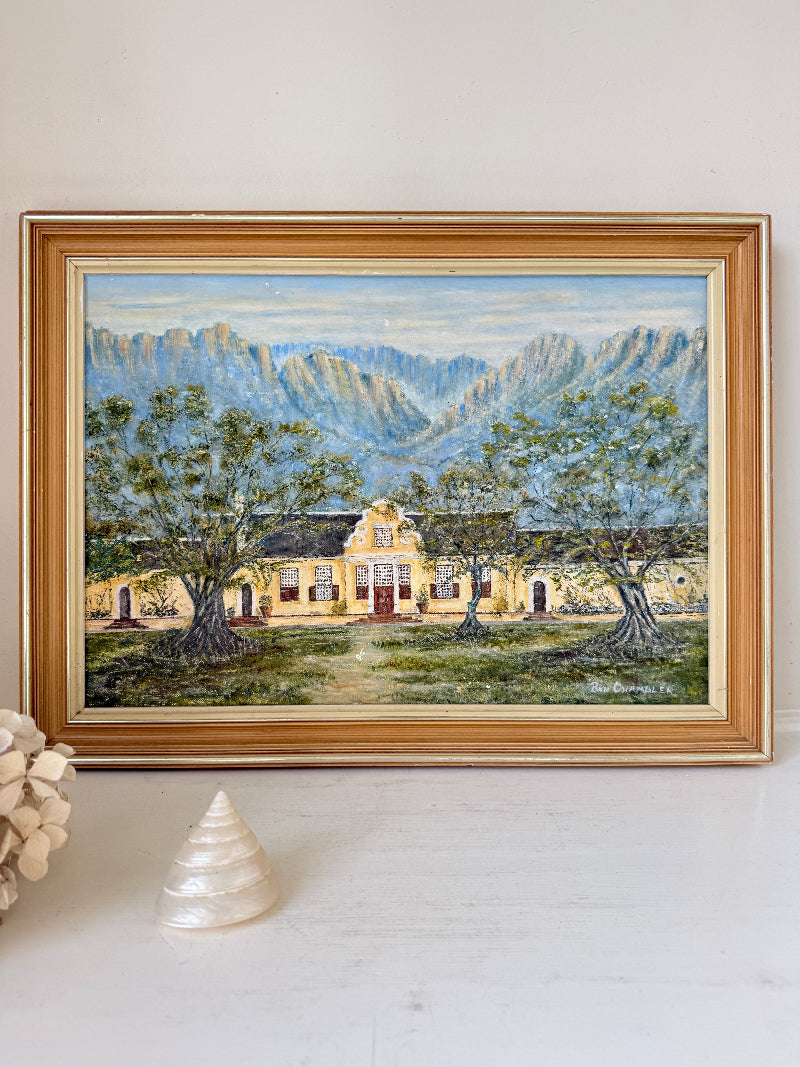 A framed landscape painting of a yellow building set among green trees with blue-gray mountains in the background, displayed on a white surface with a decorative shell and dried flowers nearby.
