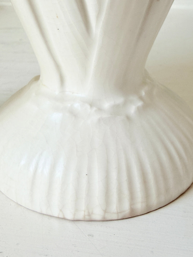 Close-up of a white ceramic vase base with a fluted, ribbed design and a narrow neck, set against a light off-white background.