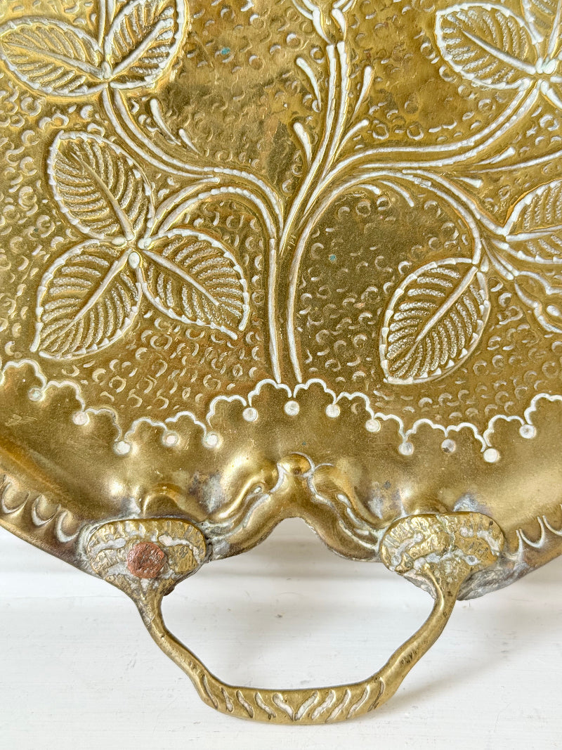 Vintage brass tray with embossed floral and leaf design, scalloped edge, and twin handles, photographed on a white surface