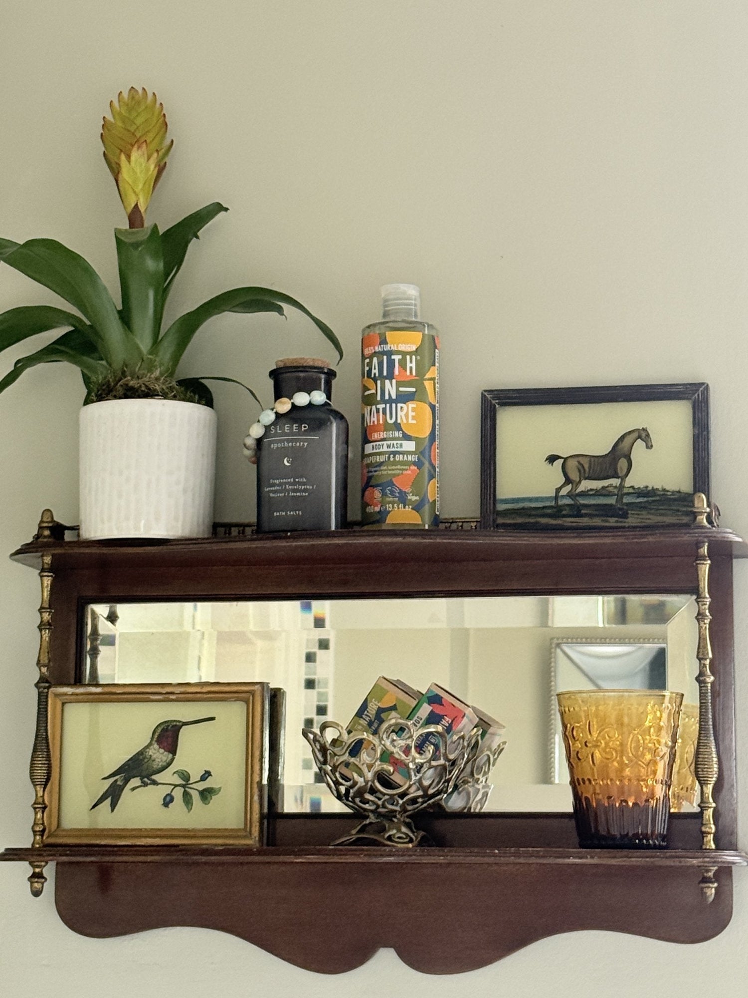 A vintage mahogany mirror shelf styled warm and cosy, with amber pressed glass, framed reverse-glass paintings of a horse and a hummingbird, a brass basket and a leafy bromeliad