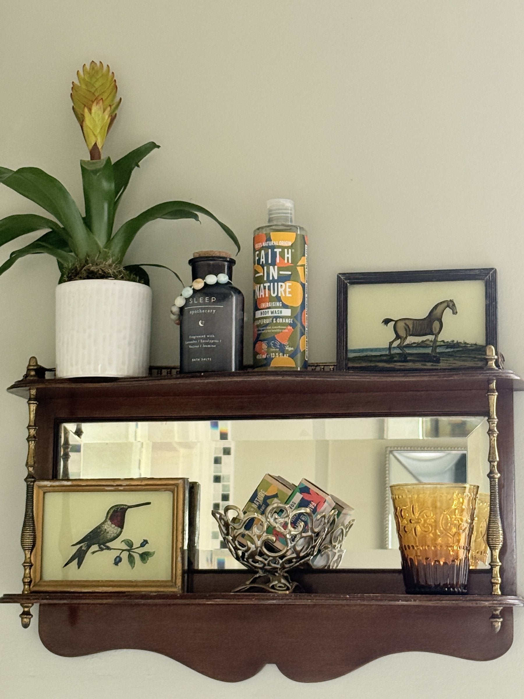 A vintage mahogany mirror shelf styled warm and cosy, with amber pressed glass, framed reverse-glass paintings of a horse and a hummingbird, a brass basket and a leafy bromeliad