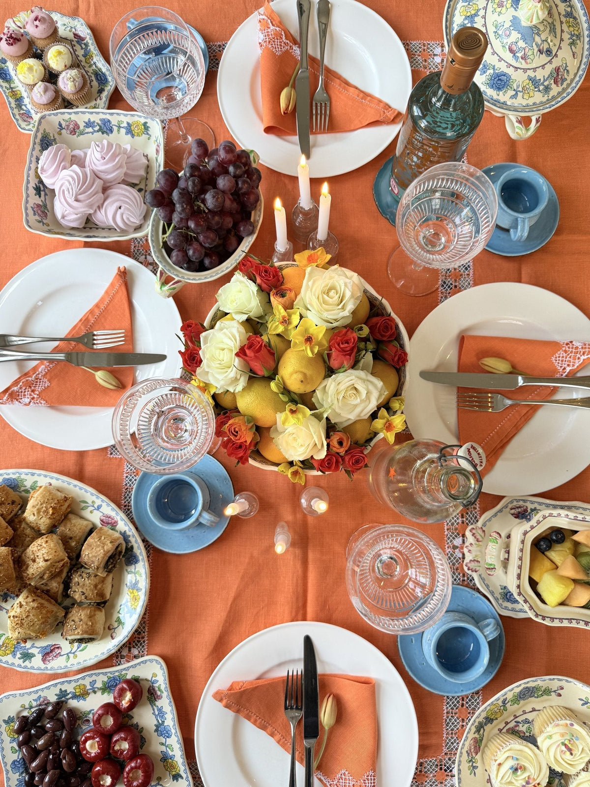 An overhead view of a summer table dressed with a bright orange linen cloth, white plates, cornflower blue coffee cups and a centrepiece of pale roses with whole lemons tucked among the blooms
