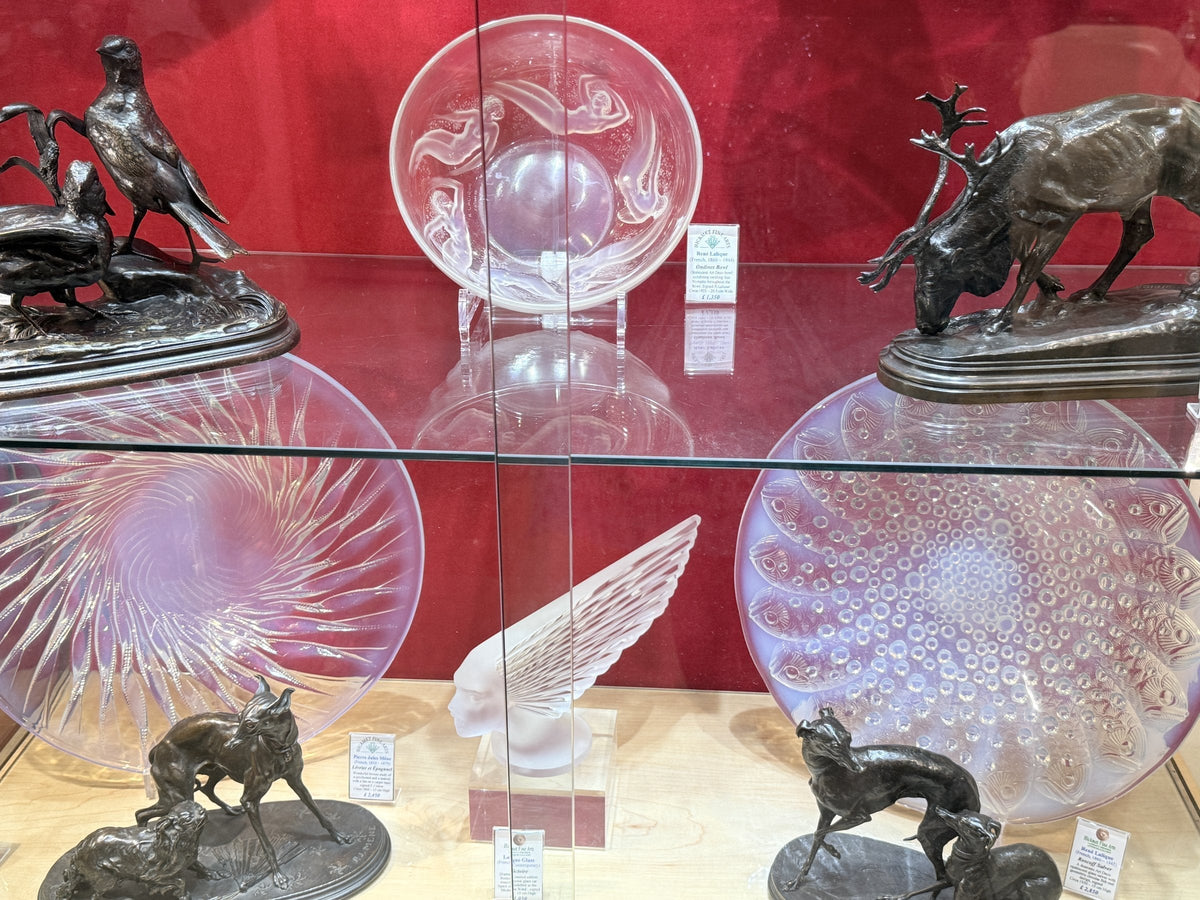 A dealer's shelf of opalescent Lalique dishes, three moulded chargers and a frosted winged bird figure arranged between small bronze animal sculptures against red velvet