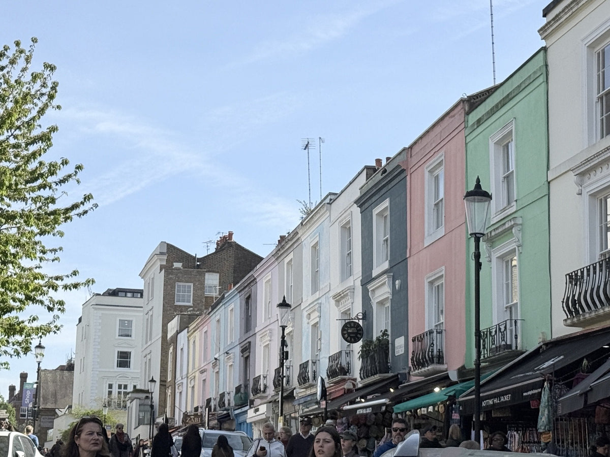 Looking up Portobello Road on a bright day, a run of pastel painted shop fronts with awnings and a busy crowd of browsers