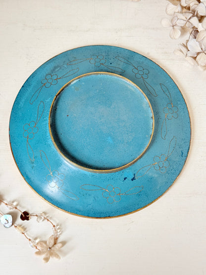 A round turquoise blue ceramic plate with a distressed finish and delicate gold floral engravings, displayed on a light cream wooden surface with decorative dried flowers.