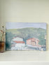 Unframed vintage oil painting of hillside cottages with terracotta roofs, green landscape, and textured brushwork, on white surface.