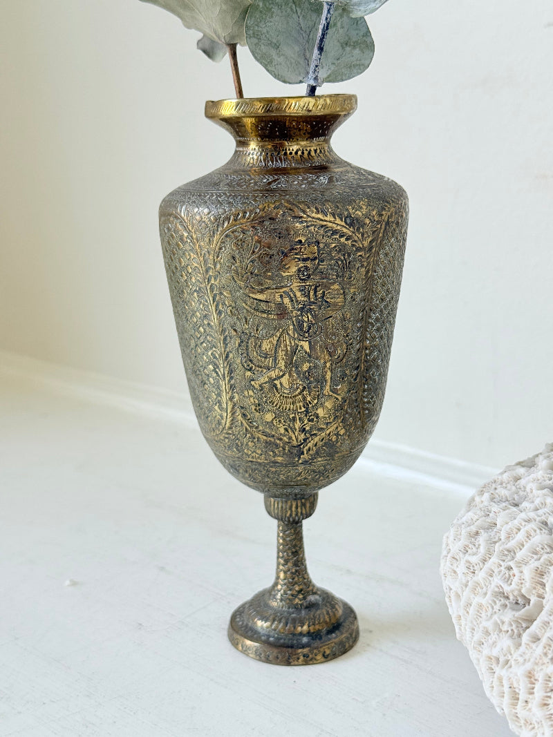 Indian Benares brass chalice vase with figural panels and pedestal base, photographed on a white surface