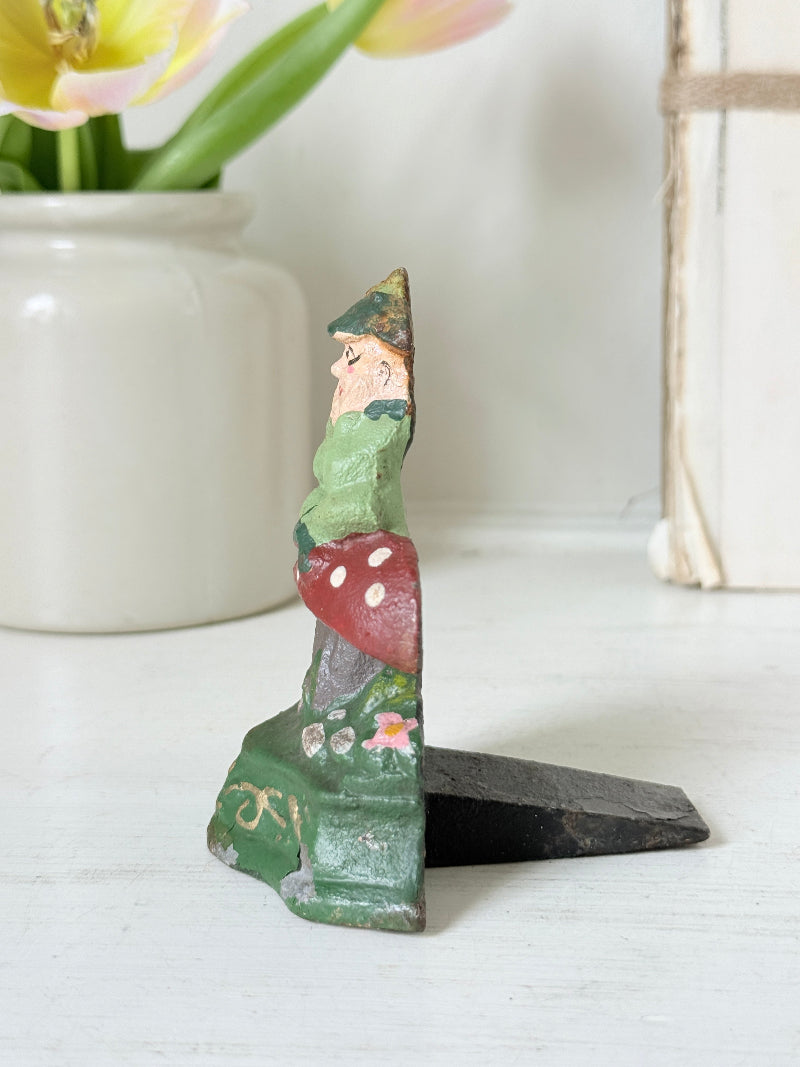 A small hand-painted wooden gnome figurine standing on a black base, displayed on a white tabletop with a white ceramic vase of tulips in the background.