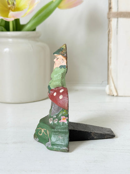 A small hand-painted wooden gnome figurine standing on a black base, displayed on a white tabletop with a white ceramic vase of tulips in the background.