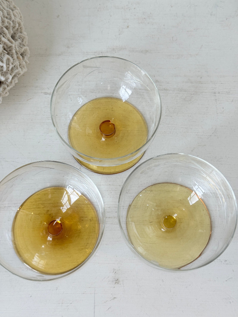 Three clear glass goblets with a circular amber-colored center, arranged on a white wooden surface.
