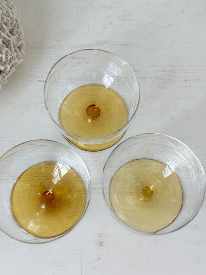 Three clear glass goblets with a circular amber-colored center, arranged on a white wooden surface.