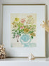 A framed botanical art print featuring a blue patterned vase filled with colorful flowers, displayed on a white surface with decorative dried grass and a shell nearby.