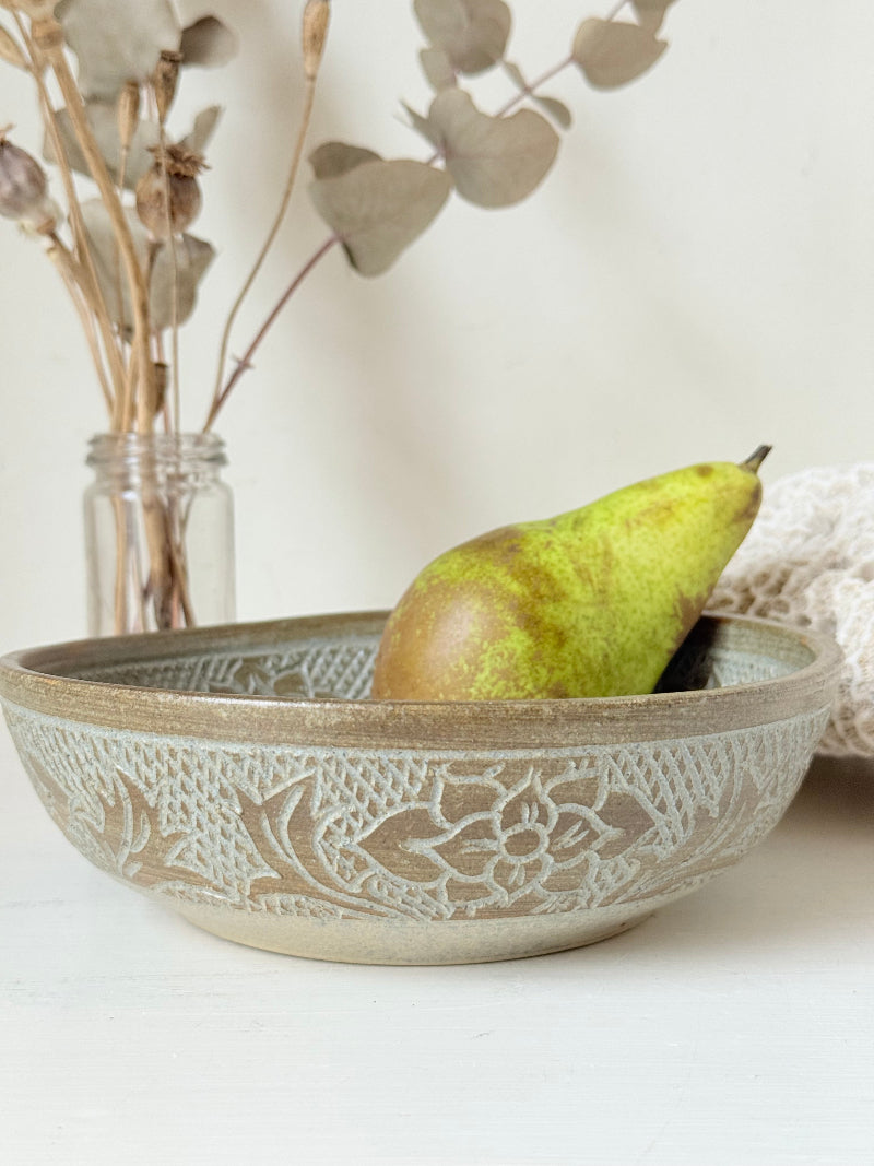 Studio pottery bowl with carved floral and geometric decoration in neutral glaze, photographed on a white surface with a pear inside.