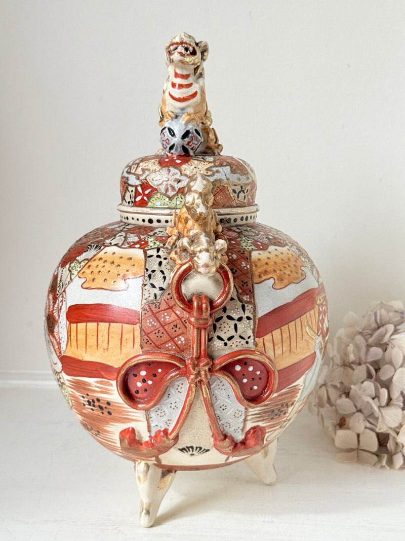 An ornate Japanese Imari-style ceramic lidded jar with a tiger figurine on top, featuring hand-painted red, gold, and white patterns on a white background.